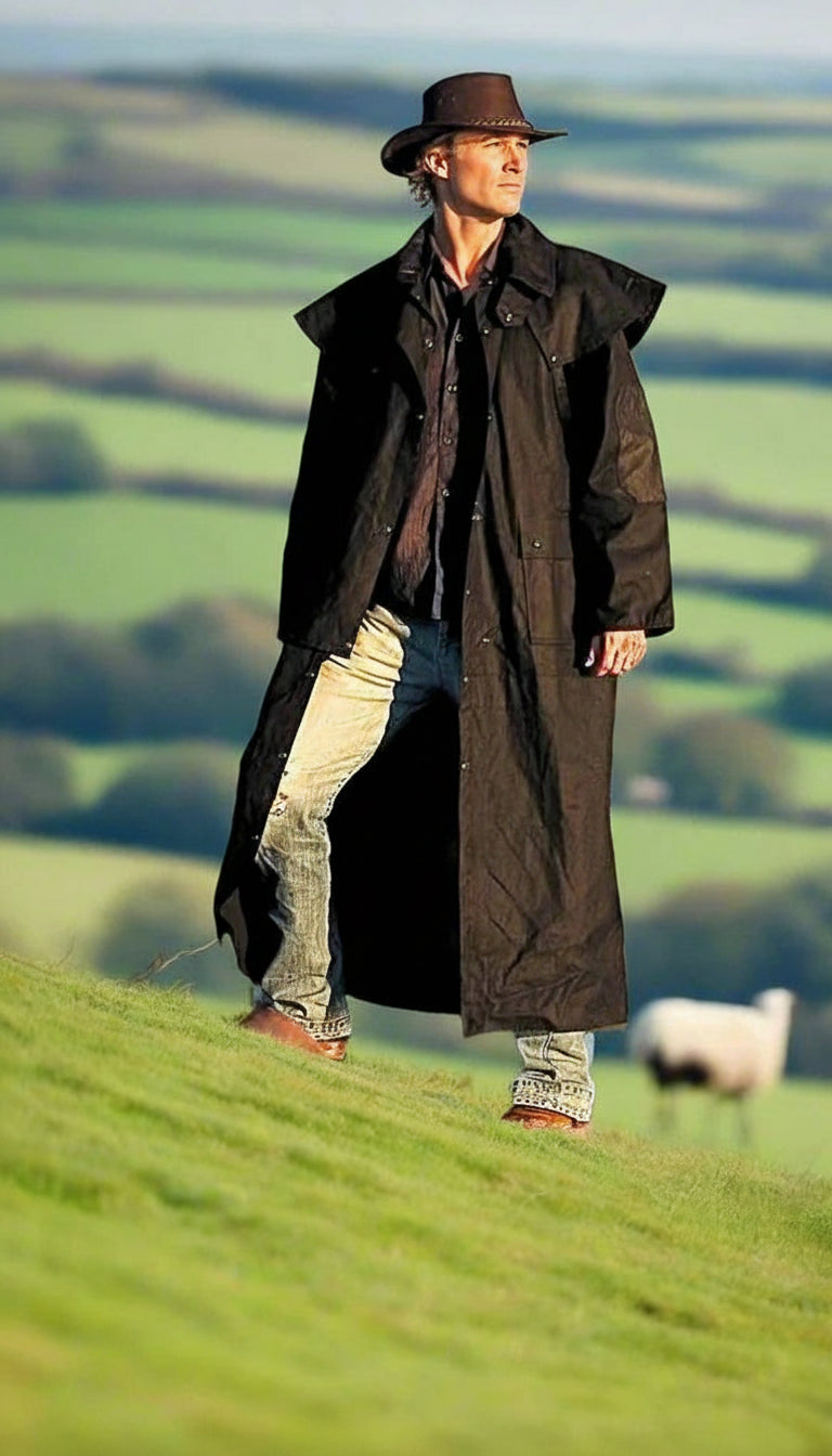 Man in a long black coat and hat standing on a grassy hill with a sheep in the background.